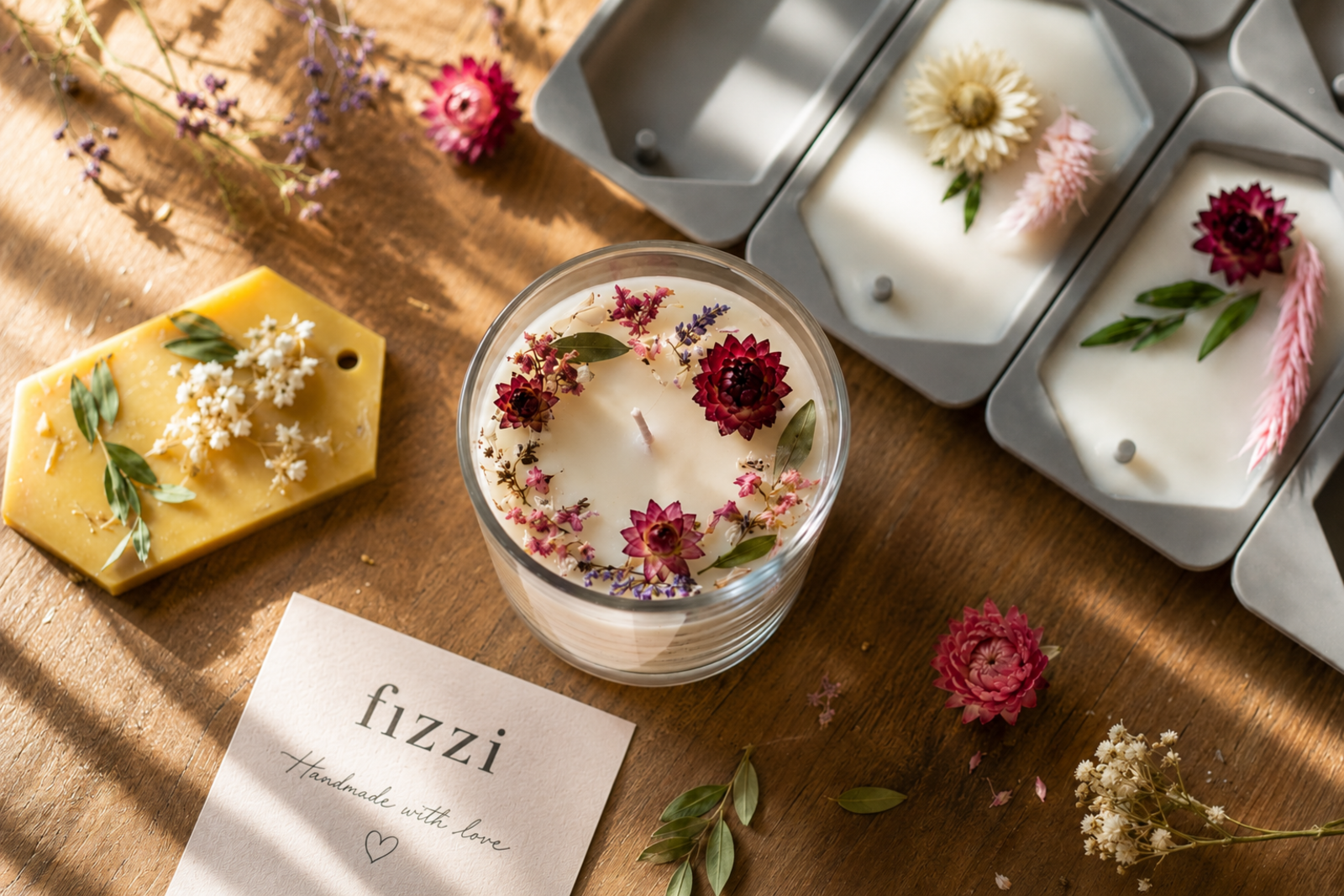A botanical-decorated soy candle, beeswax soap bar, and bath salt molds with dried flowers arranged on a warm wooden table, alongside a card reading 'fizzi — handmade with love' in dappled afternoon light.